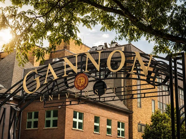 The gold GANNON UNIVERSITY lettering on a black iron arch, brick campus buildings behind it and trees overhead.